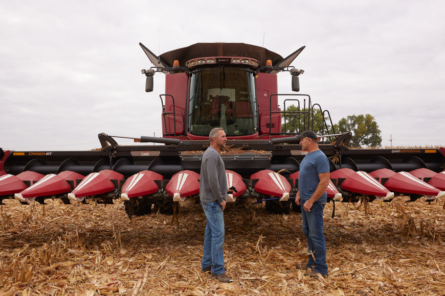 Two farmers talking in front of a combine in a harvested corn field.