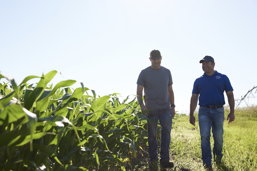 A grower and a retailer walk side by side smiling next to a corn field. 