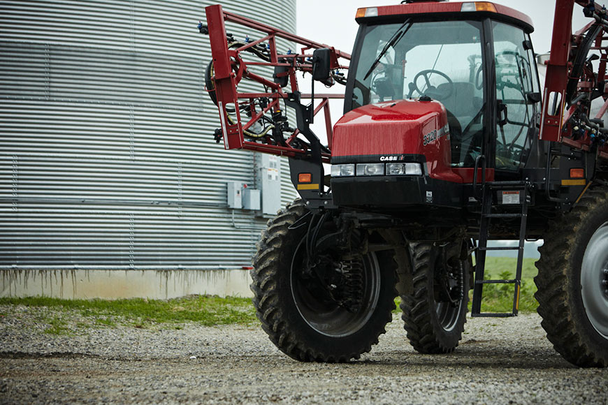 Red sprayer parked on farm yard in front of silver grain bin